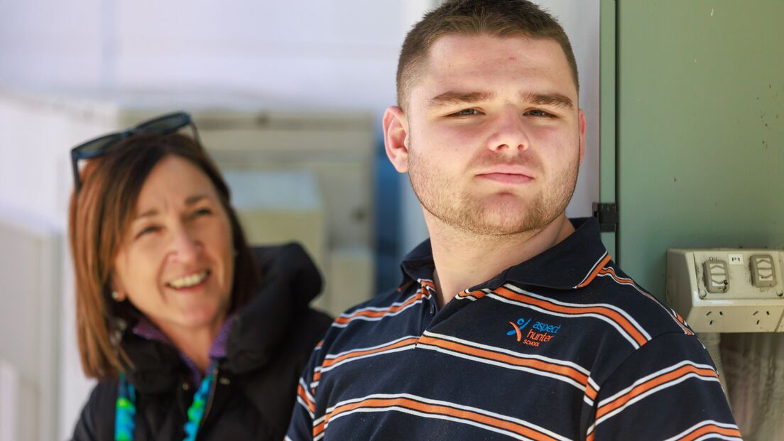 A young man with short hair and a trimmed beard stands outside, looking off into the distance with a thoughtful expression. He is wearing a navy blue, orange, and white striped polo shirt with a logo that reads "Aspect Hunter School." Behind him, a woman with shoulder-length brown hair smiles warmly in his direction. She is dressed in a black jacket and has sunglasses resting on her head. The setting appears to be outdoors, with soft, natural lighting and a relaxed atmosphere.